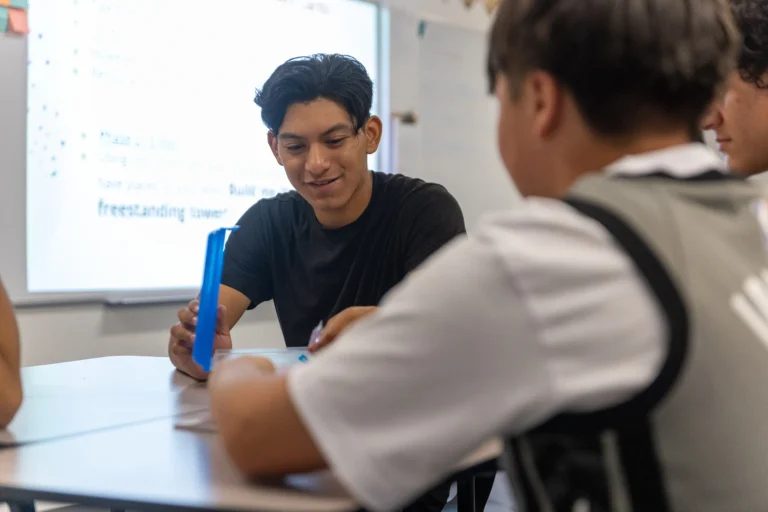 A teenage boy in a black t-shirt smiles while holding a blue object and sitting at a table with two other students in a classroom. A whiteboard with text is visible in the background.