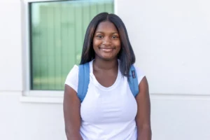 A young woman wearing a white t-shirt and blue backpack stands in front of a window, smiling at the camera. The background is a light-colored building.