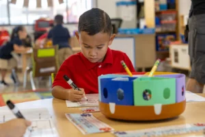 A young boy wearing a red shirt sits at a classroom table, focused on writing with a marker. A colorful round pencil holder with compartments and pencils is on the table in front of him.
