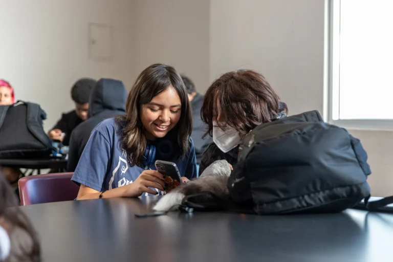 Two students sit at a desk, one smiles while showing something on her phone to the other, who wears a white face mask. Backpacks and more students are in the background near a sunlit window.