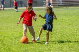 Two young children play soccer on a grassy field. The boy in a red shirt and glasses tries to kick an orange ball while the girl in a blue dress and ponytail moves to block him.
