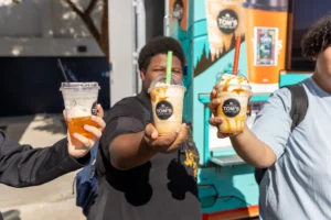 Three people hold up iced beverages in clear cups labeled “Tom’s” in front of a colorful food truck, with two drinks topped with caramel drizzle and whipped cream. The background is outdoors and sunny.