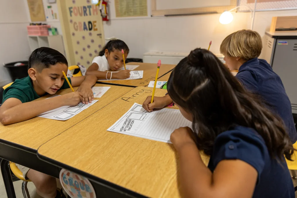 Four elementary school children sit at a table, focused on writing assignments with pencils in a classroom labeled Fourth Grade in the background.