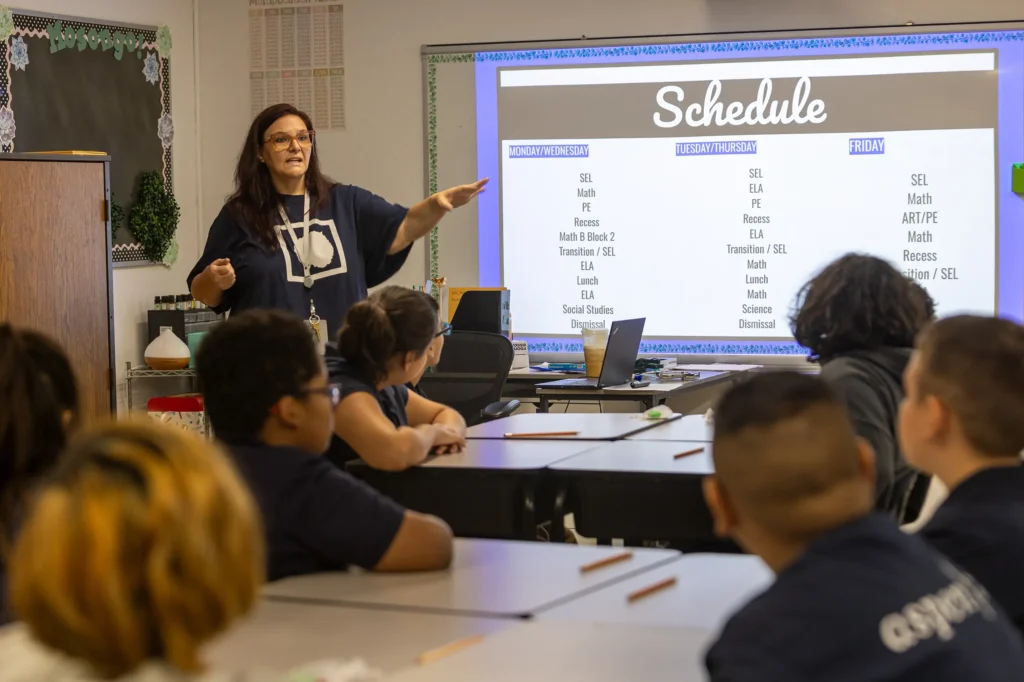 A teacher stands at the front of a classroom, pointing to a large schedule projected on a screen while students seated at desks listen attentively. The schedule lists subjects for Monday, Tuesday/Thursday, and Friday.