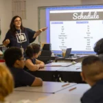 A teacher stands at the front of a classroom, pointing to a large schedule projected on a screen while students seated at desks listen attentively. The schedule lists subjects for Monday, Tuesday/Thursday, and Friday.