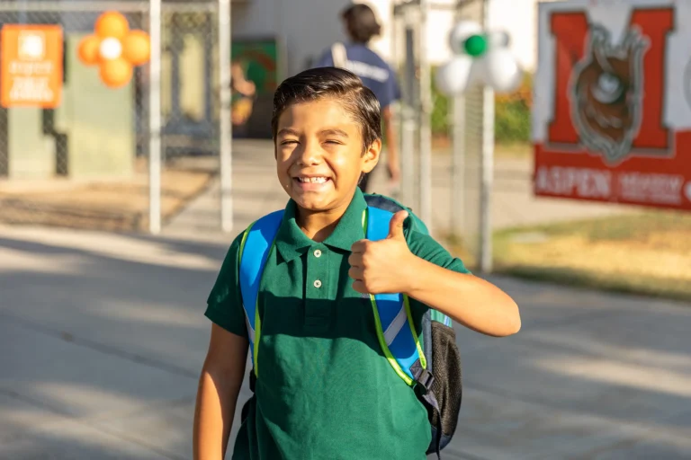 Smiling young boy with a backpack gives a thumbs-up outside a school, with sunlight and colorful balloons in the background.