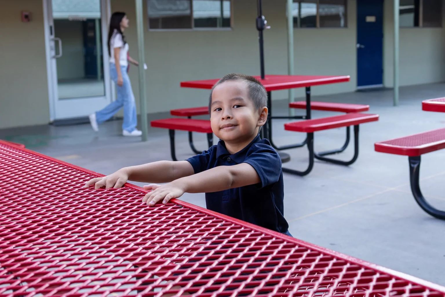 A young boy stands outdoors, holding onto a red metal picnic table. He wears a dark shirt and smiles slightly. Behind him, there are more red tables, a woman walking, and a building with doors and windows.