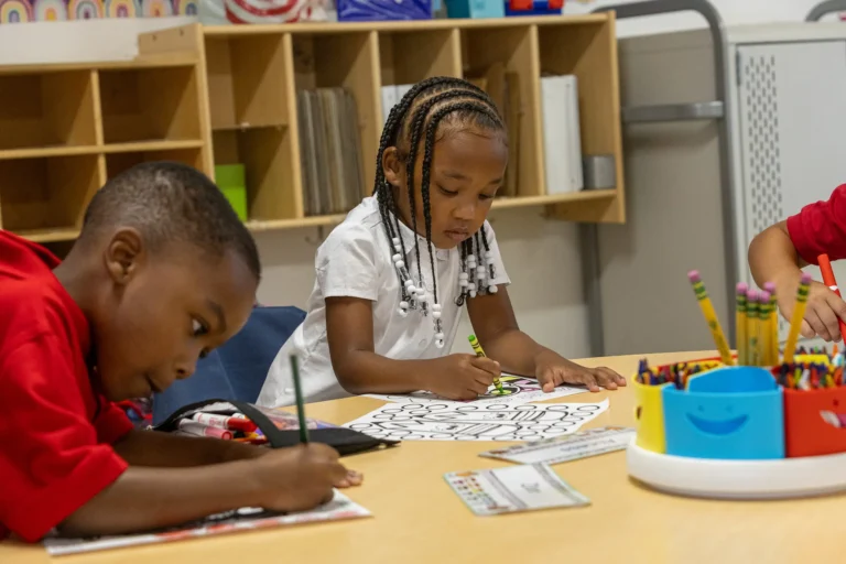 Two young children sit at a classroom table, coloring worksheets with crayons and pencils. Shelves and supplies are in the background, and a container of colorful pencils sits on the table.