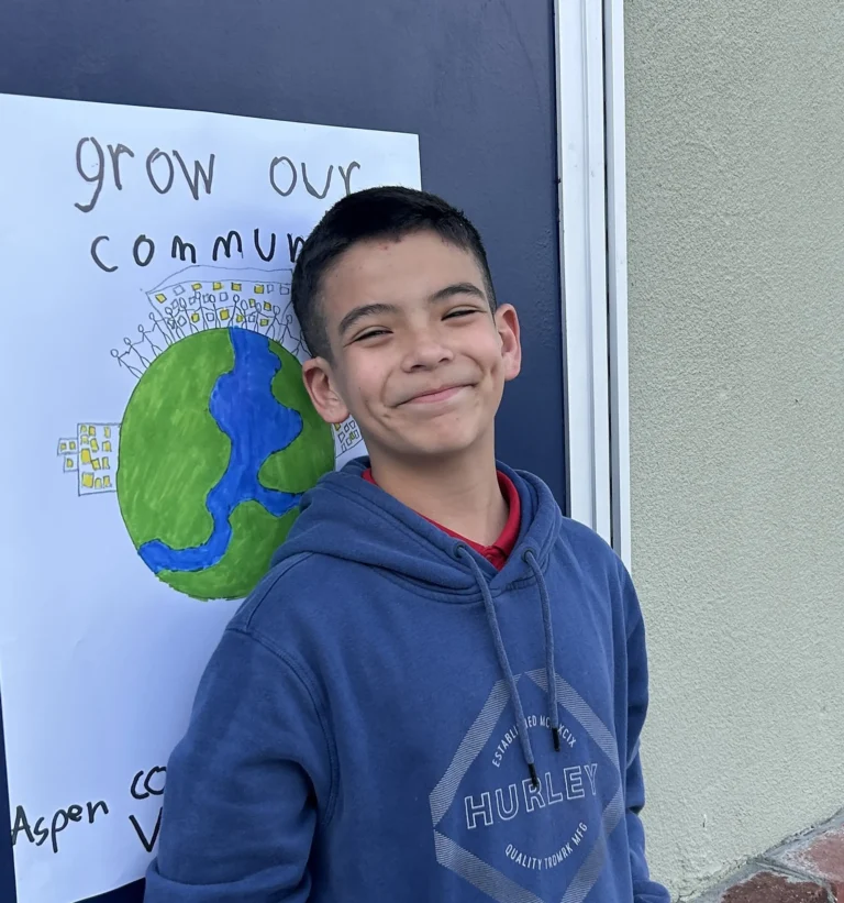A smiling boy in a blue Hurley hoodie stands in front of a poster that reads grow our community and shows a drawing of the Earth with people holding hands around it.