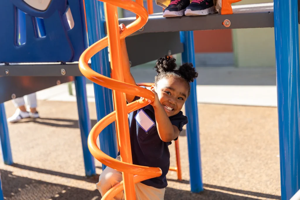 A young girl with pigtails smiles while climbing an orange spiral ladder on a playground structure, with sunlight illuminating the scene.