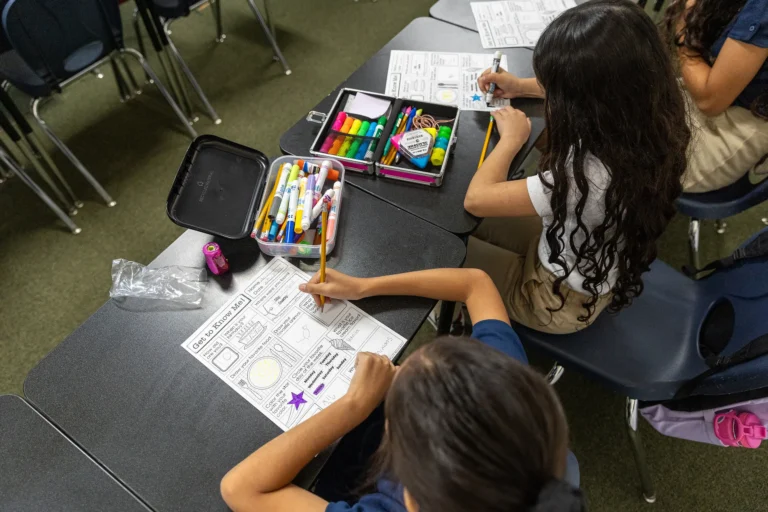 Two students sit at desks working on worksheets with drawings and text. Art supplies like markers, crayons, and a pencil case are on the desks. The students have long hair and are seen from above in a classroom setting.