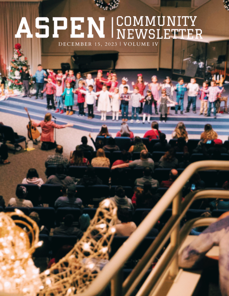 Children stand in a row on stage performing for an audience in a decorated auditorium with a Christmas tree, as seen on the cover of Aspen Community Newsletter, December 15, 2023, Volume IV.