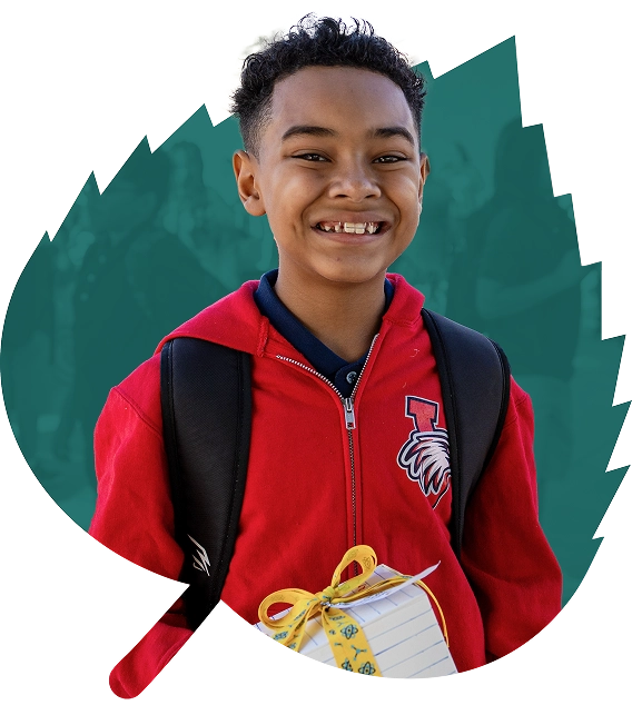 Smiling boy in a red jacket with a school emblem, wearing a backpack, holds a yellow-ribbon-wrapped gift against a teal, leaf-shaped background.