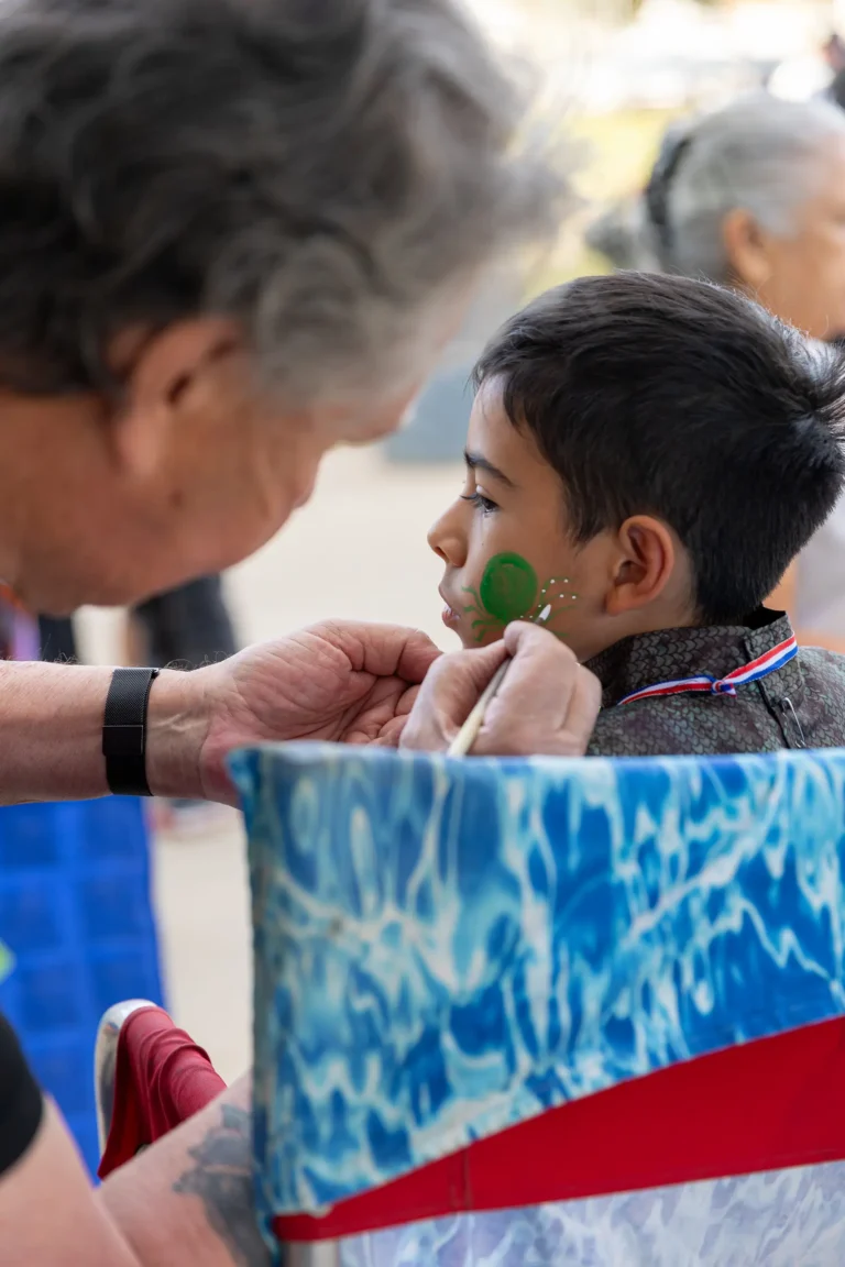 An artist paints a green design with white dots on a young boy’s cheek. The boy sits still in a chair with a blue patterned cover, while an older person carefully applies face paint with a small brush.