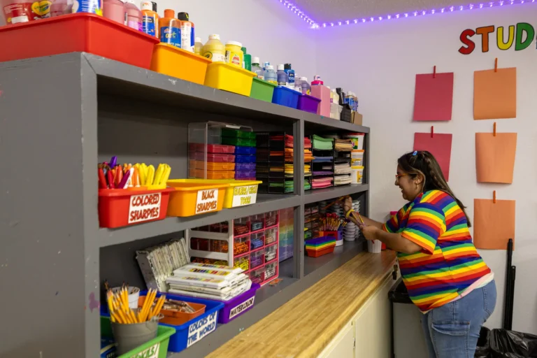 A woman in a rainbow-striped shirt organizes colorful art supplies on shelves in a classroom. The shelves hold markers, paper, glue, and paints. Blank sheets of paper hang on the wall in the background.