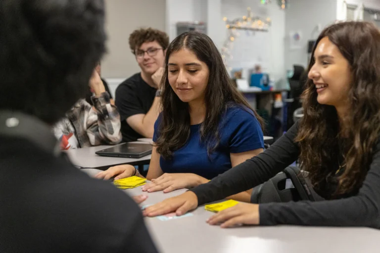 A group of students sit around a table in a classroom, smiling and interacting while playing a card game. Two girls are in focus, one wearing a blue shirt and another in black, both looking at cards on the table.