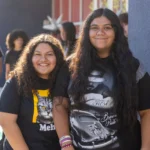 Two teenage girls with long dark hair smile at the camera. One wears a shirt with a cat graphic, the other wears a shirt with a person in a hat. Other people are blurred in the background outdoors.