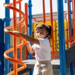 A young girl smiles while holding onto a bright orange spiral climbing bar at a colorful playground, with various blue and yellow play structures in the background.