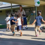 A group of young children in school uniforms run outside near a covered walkway, with a teacher watching and colorful balloons hanging overhead.