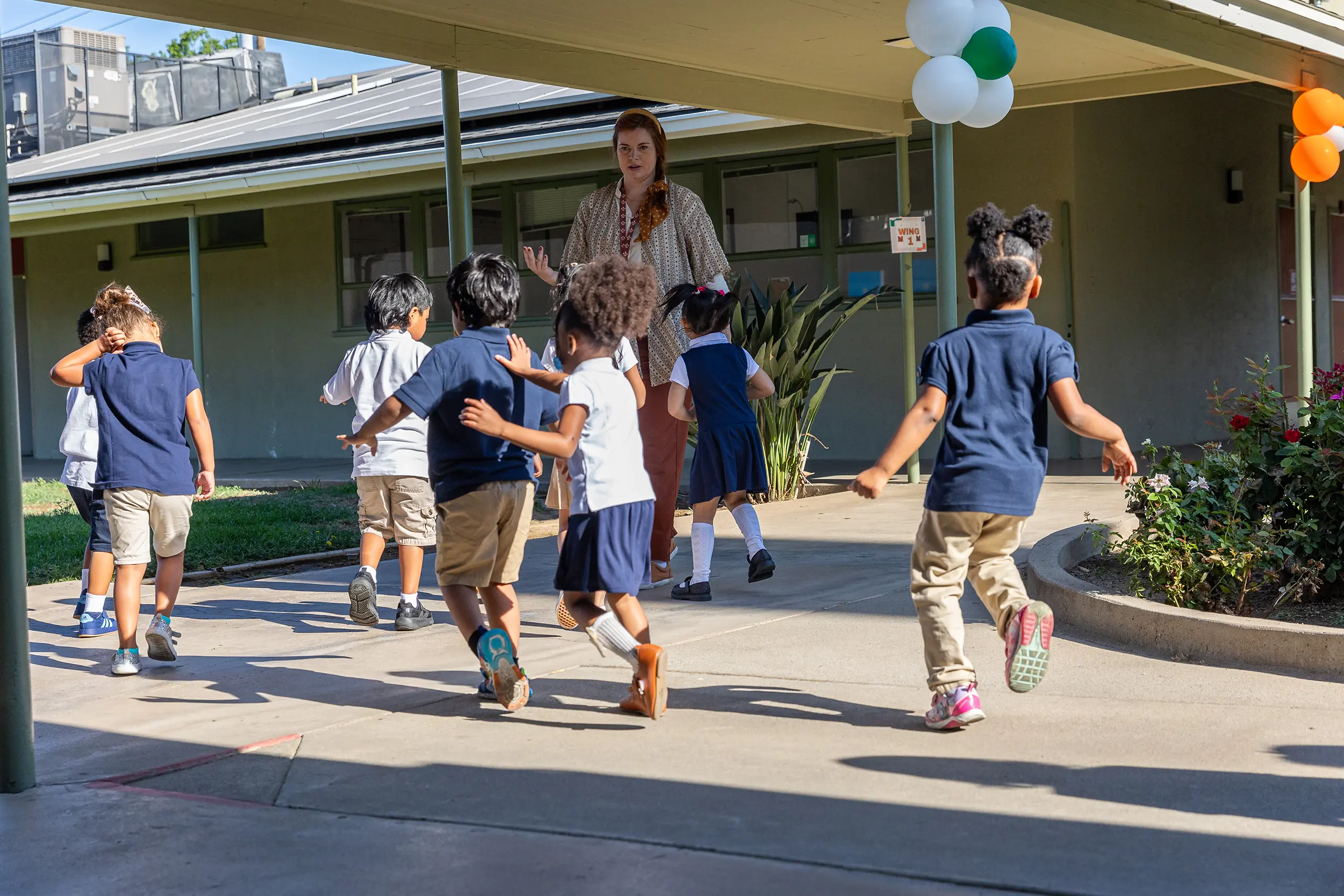 A group of young children in school uniforms run outside near a covered walkway, with a teacher watching and colorful balloons hanging overhead.