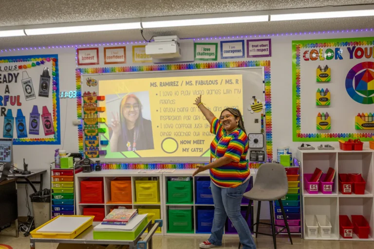 A teacher in a colorful classroom points and smiles at a screen showing her photo and fun facts about herself. The room is decorated with bright bins, posters, and educational materials.