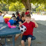 A group of children sit at a picnic table outdoors, some eating lunch and waving. One boy in a red shirt sits on the bench holding a soccer ball and smiling. There is a tree providing shade and a playground in the background.