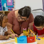 A teacher helps two young children with a coloring activity at a classroom table, surrounded by pencils, markers, and art supplies.